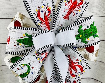 May include: A white, black, and red Christmas bow with a white center. The bow is made of ribbon with a woodgrain pattern and features images of alligators, lobsters, and Christmas lights.