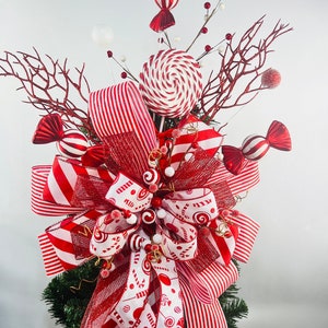 May include: A festive Christmas tree topper featuring red and white striped ribbons, candy-shaped ornaments, and a large swirl lollipop. The topper is decorated with red berries and branches, creating a cheerful holiday display.