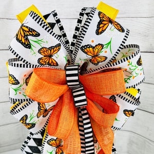 May include: A large orange bow with a black and white checkered ribbon and a white ribbon with a butterfly print. The bow is tied with a long orange ribbon tail.