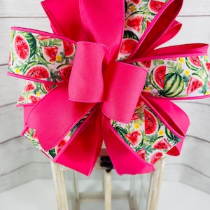 May include: A large, bright pink bow with a watermelon-themed ribbon. The ribbon features a white background with red watermelon slices, green watermelons, and yellow flowers. The bow is likely for decoration.