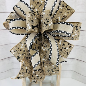 May include: A decorative bow featuring a tan and black dog bone and paw print pattern. The bow has a burlap texture with black and white polka dot trim. The bow is a decorative accent for gifts or home decor.