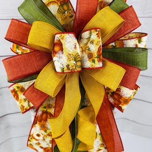 May include: A large, multi-layered bow made with burlap ribbon in shades of green, orange, and yellow. The center of the bow features a printed ribbon with a fall-themed pattern of pumpkins and sunflowers.