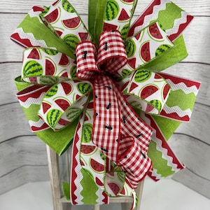 May include: A decorative bow featuring watermelon slices and green and red gingham patterns. The bow has multiple layers of ribbon in green, red, and white, with a central knot. The bow is placed on a white wooden lantern.