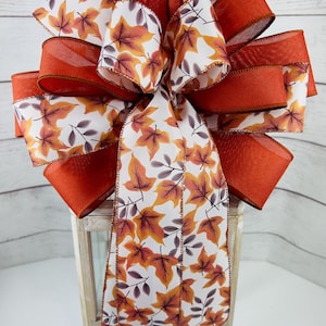 May include: A decorative bow featuring a fall leaf pattern. The bow has a combination of solid orange and leaf-printed ribbons. The leaf pattern includes orange, yellow, and brown leaves on a white background. The bow is likely for seasonal decor.