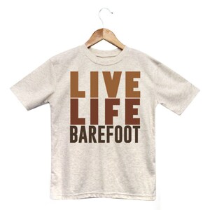 以下が含まれることがあります： 「LIVE LIFE BAREFOOT」の文字が茶色でプリントされた、ライトグレーのTシャツ。文字は縦に積み重ねられています。Tシャツは白い背景に、木製のハンガーに掛けられています。