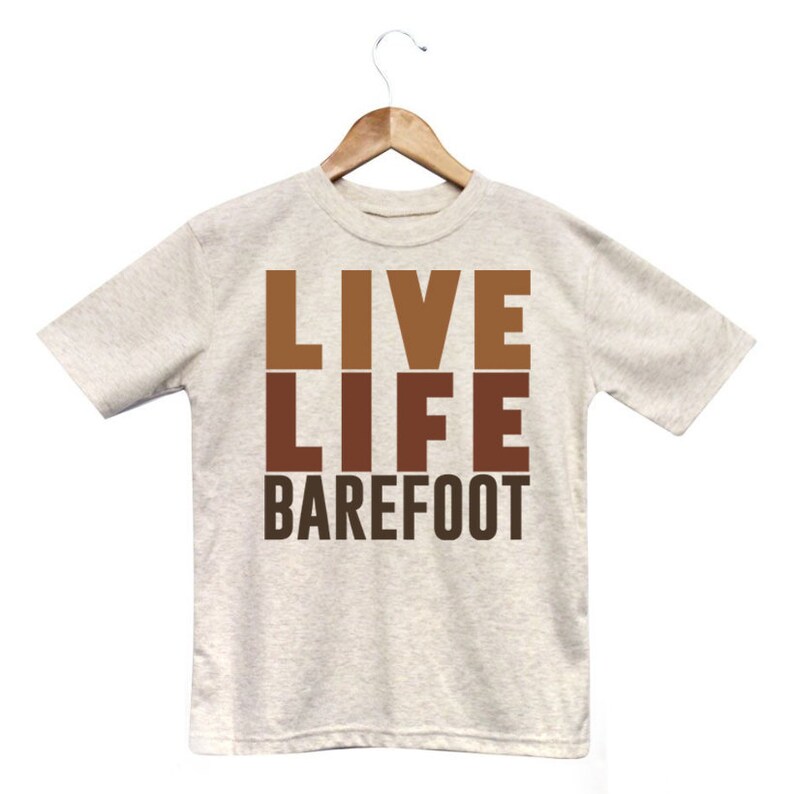 以下が含まれることがあります： 「LIVE LIFE BAREFOOT」の文字が茶色でプリントされた、ライトグレーのTシャツ。文字は縦に積み重ねられています。Tシャツは白い背景に、木製のハンガーに掛けられています。