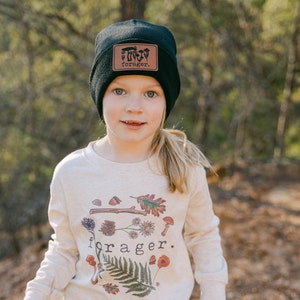 May include: A young person wearing a black beanie with a brown leather patch that says "forager." The beanie has a small illustration of mushrooms on the patch. The person is wearing a cream colored long-sleeved shirt with a graphic of mushrooms, leaves, and flowers. The shirt says "forager."