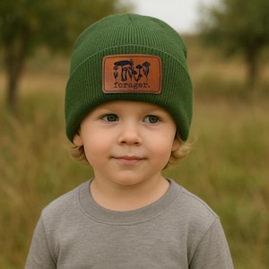 May include: A green beanie with a brown leather patch featuring a mushroom design and the word "forager." The beanie is made of a ribbed knit material. The child is wearing a gray long-sleeve shirt.