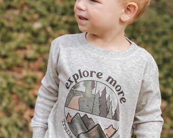 May include: A gray long-sleeve sweatshirt with a circular graphic that says "explore more" and depicts a path through a forest, mountains, and the words "trails", "forests", and "mountains".