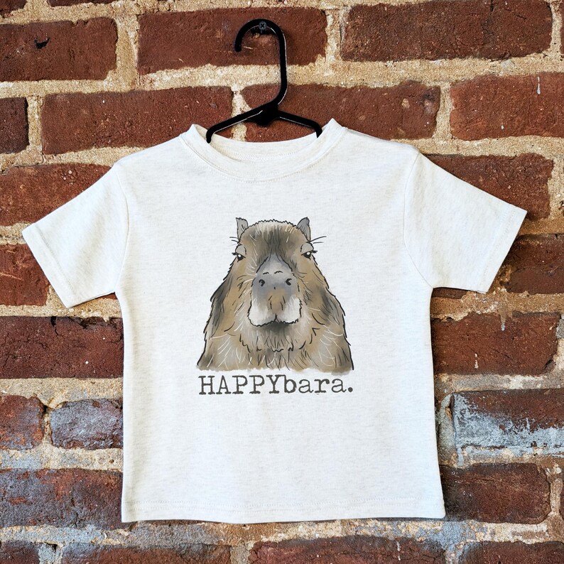 May include: A light gray t-shirt featuring a detailed illustration of a capybara's face. The capybara is depicted in shades of brown and gray. Below the image is the text "HAPPYbara."