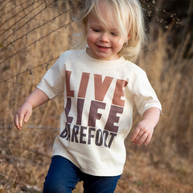 以下が含まれることがあります： 茶色と濃い茶色のブロック文字で「LIVE LIFE BAREFOOT」と書かれたベージュのTシャツを着た子供。子供は青いジーンズを履いていて、笑顔です。背景は、背の高い乾燥した草でぼやけています。