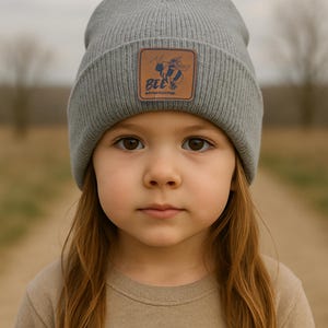 May include: A gray beanie with a brown leather patch featuring a cartoon bee and the text "BEE'S adventurous." The beanie is worn by a young child with long brown hair and a tan shirt.