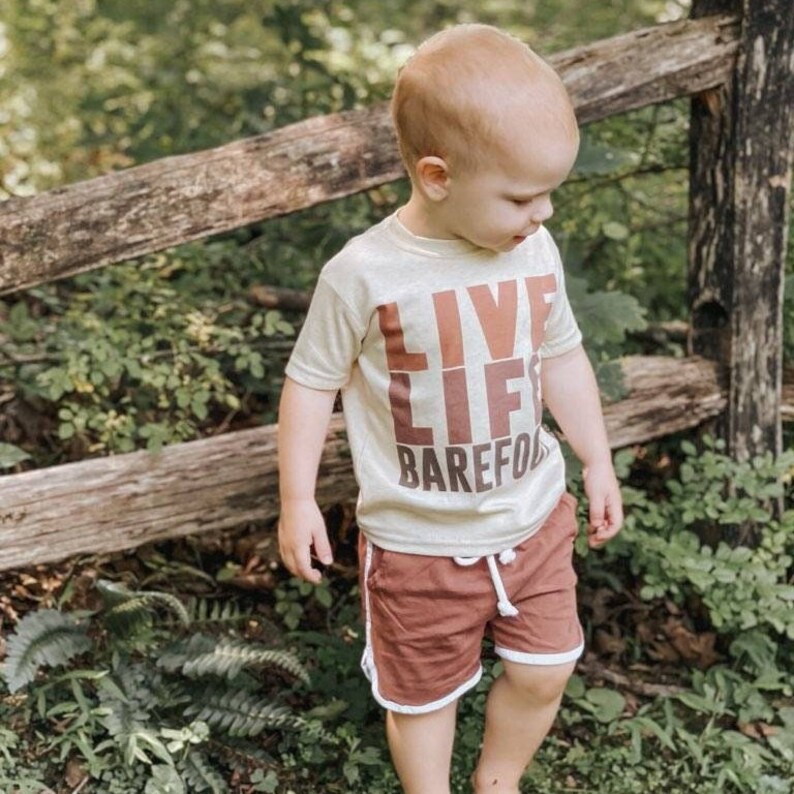 以下が含まれることがあります： 「LIVE LIFE BAREFOOT」の文字が茶色でプリントされたベージュ色のTシャツを着た子供。白いトリムが付いた錆色のショーツも着用しています。背景には木の柵と緑があります。