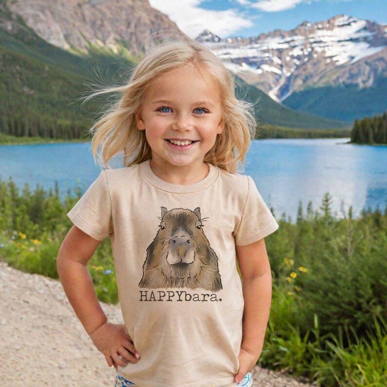 May include: A young child wearing a tan t-shirt with a graphic of a capybara and the text "HAPPYbara." The child is smiling in front of a lake and mountain backdrop.