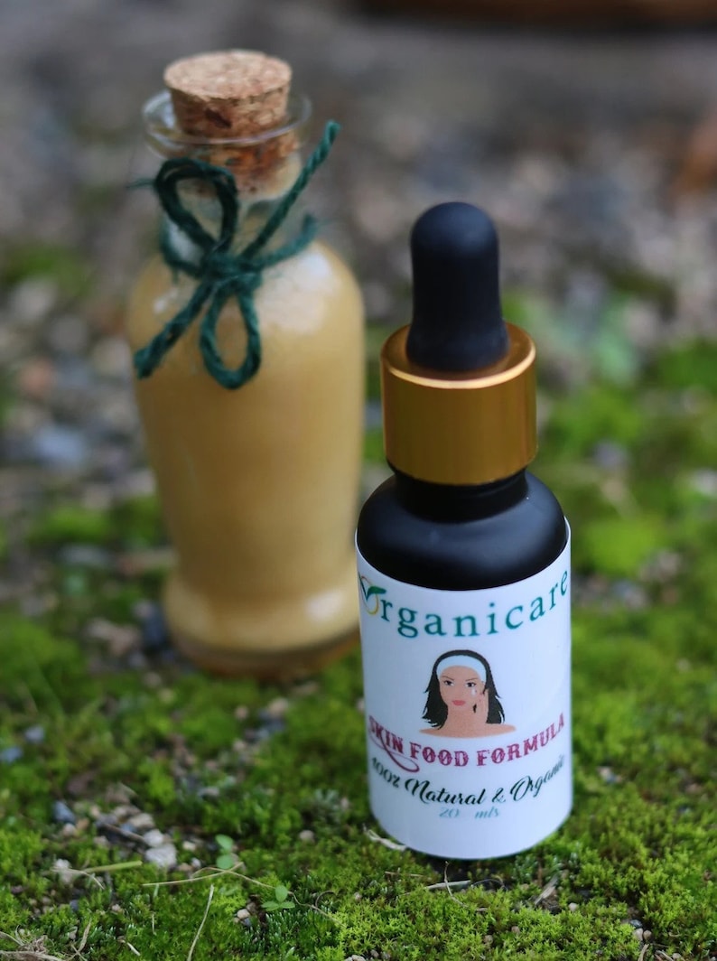 May include: Two glass bottles with labels. The smaller bottle is a dark amber color with a gold lid and a dropper. The label says "Organicare Skin Food Formula 100% Natural & Organic 20 ml". The larger bottle is clear with a cork stopper and a green ribbon. The background is green moss.