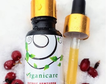 May include: A glass bottle of organic hawthorn face and body oil with a gold lid and dropper. The bottle is sitting on a white surface with red berries scattered around it.