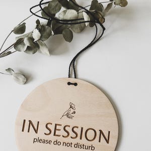May include: A round wooden sign with a black string for hanging. The sign reads "IN SESSION please do not disturb" and features a small floral design.