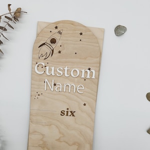 May include: A wooden growth chart with a rocket ship and stars design. The chart has a space for a custom name and numbers to track height. The text "six" is visible on the chart.