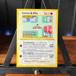 May include: A wedding invitation card displayed on a small black easel. The card features a colorful, pixelated graphic with the names "James & Ella" and the date "April 16, 2026." Text on the card details the ceremony and reception information.