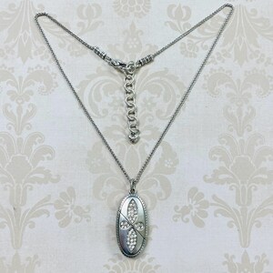 May include: Silver-toned necklace with an oval pendant. The pendant features a cross design encrusted with clear crystals. The necklace has a delicate chain and an adjustable clasp. The background has a floral pattern.