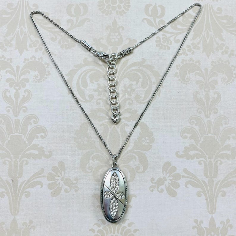 May include: Silver-toned necklace with an oval pendant. The pendant features a cross design encrusted with clear crystals. The necklace has a delicate chain and an adjustable clasp. The background has a floral pattern.