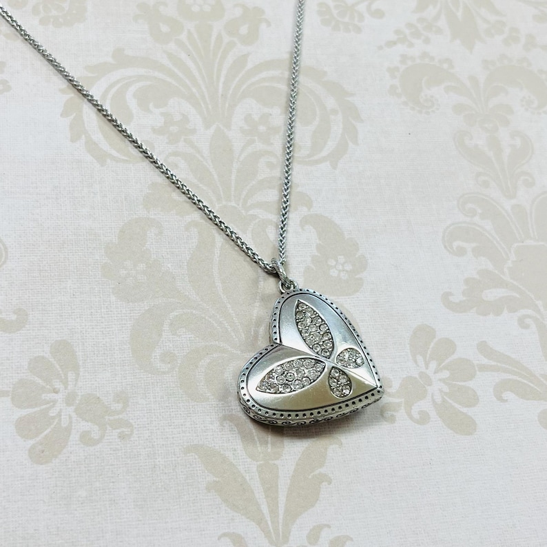 May include: Silver-toned heart pendant necklace. The heart-shaped pendant features a butterfly design with clear crystals and a beaded border. The necklace chain is a delicate silver color.