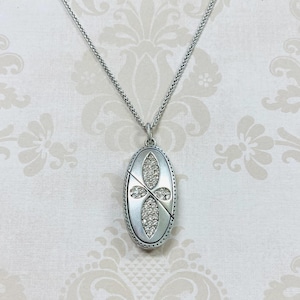 May include: Silver-toned necklace with an oval pendant. The pendant features a cross design with clear, sparkling stones. The necklace has a delicate chain and is set against a patterned background.