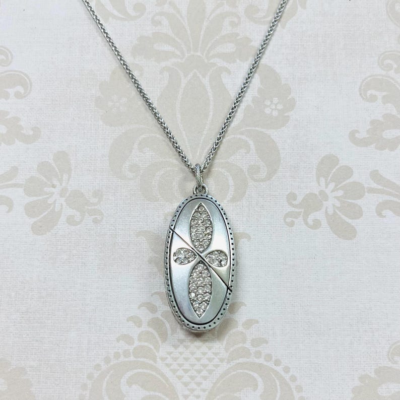 May include: Silver-toned necklace with an oval pendant. The pendant features a cross design with clear, sparkling stones. The necklace has a delicate chain and is set against a patterned background.
