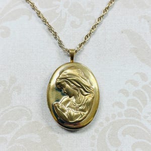 May include: A gold-toned oval locket pendant suspended from a matching twisted rope chain. The front of the locket features a raised, embossed relief depicting a profile of a figure with long hair, wearing a garment with a small cross necklace.
