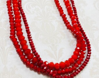 May include: A multi-strand necklace featuring faceted red beads of varying sizes. The necklace has a layered design, with strands of different lengths and bead sizes, creating a textured and eye-catching accessory. The beads have a glossy finish.