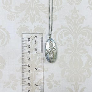 May include: Silver-toned oval pendant necklace with a cross-shaped design of clear crystals. The pendant is approximately 1.5 inches long. The necklace chain is a delicate, silver-colored chain.