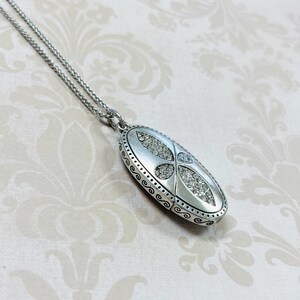 May include: Silver-toned oval pendant necklace with a decorative design. The pendant features a central design of clear crystals and is suspended from a delicate chain. The pendant is approximately 1.5 inches long.