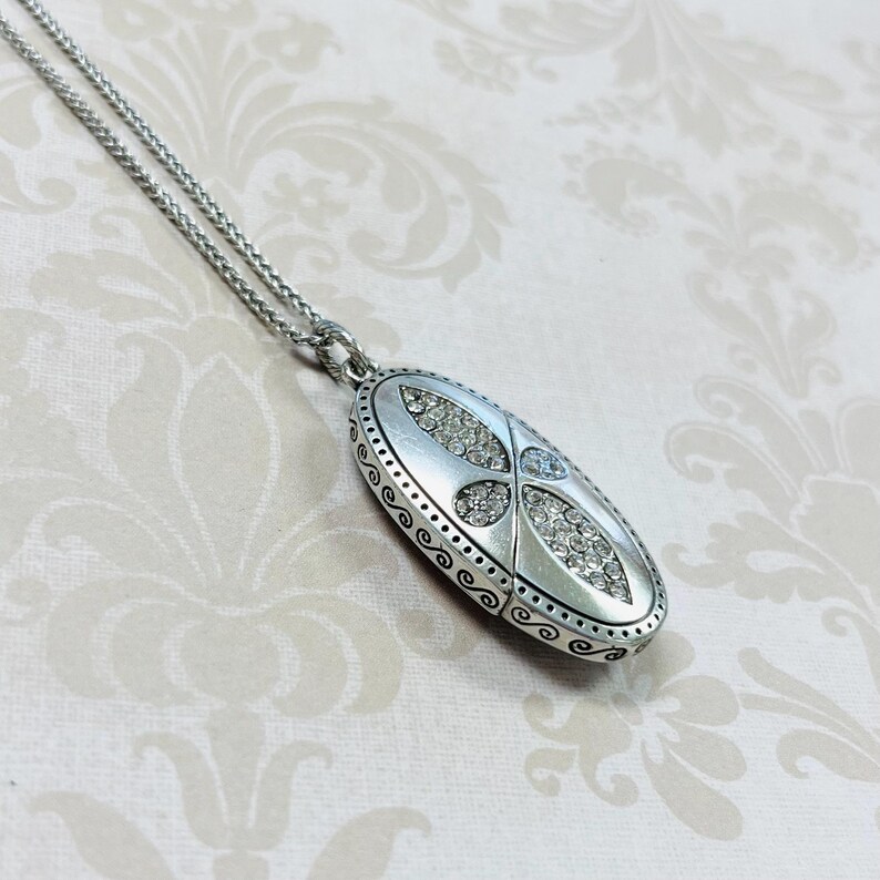 May include: Silver-toned oval pendant necklace with a decorative design. The pendant features a central design of clear crystals and is suspended from a delicate chain. The pendant is approximately 1.5 inches long.