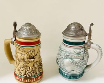 May include: Two decorative beer steins with metal lids. One stein is tan with a relief of leopards and foliage, accented with red and green bands. The other is white with a relief of marine life and green and red bands.