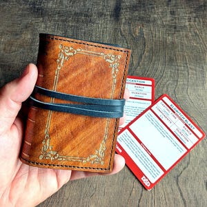 Può includere: Un portafoglio in pelle marrone con dettagli dorati e un cinturino nero. Il portafoglio è tenuto in mano, con due carte rosse e bianche visibili. Le carte hanno del testo.