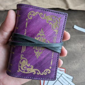 Può includere: Un piccolo diario in pelle viola con disegni floreali dorati in rilievo e un involucro in pelle nera. Il diario ha un'estetica vintage, con una finitura invecchiata sulla copertina. I bordi sono cuciti e il diario è chiuso con un cinturino in pelle.