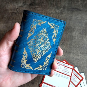 May include: A blue leather wallet with gold filigree designs. The wallet is held in a hand, with several red and white cards visible. The wallet's design is ornate and detailed, with a vintage aesthetic. The cards have text on them.
