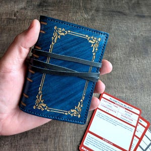 May include: A blue leather journal with gold filigree and a black leather wrap. The journal is closed and has a textured appearance. Several red and white cards are visible next to the journal.