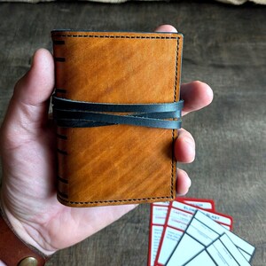 May include: A handcrafted, brown leather wallet with black stitching and a black leather strap closure. The wallet is held in a hand, showcasing its textured surface and detailed craftsmanship. The wallet appears to be new.