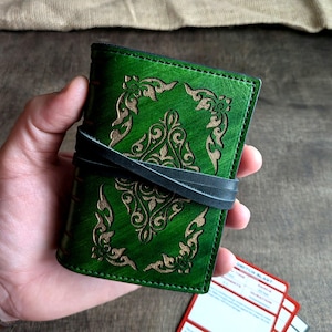 May include: A green leather journal with an embossed floral design and a black leather strap closure. The journal is held in a hand, with several game cards visible in the background. The journal's cover is a vibrant green.