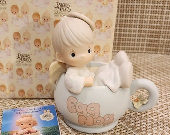 May include: A Precious Moments figurine of an angel sitting in a blue and white mug with the words "Egg Nog" on it. The figurine is part of the "Dropping In For The Holidays" collection.