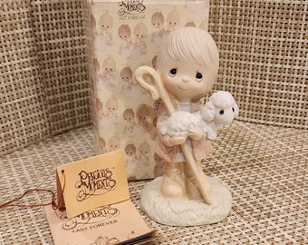 May include: A porcelain figurine of a young shepherd holding a lamb. The figurine is in a white box with a brown and white pattern. The box has the words "Precious Moments" and "Last Forever" printed on it.