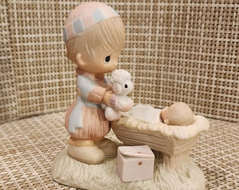 May include: A porcelain figurine of a young girl in a pink and blue dress holding a white lamb. The figurine is standing on a wooden manger with a small box next to it.