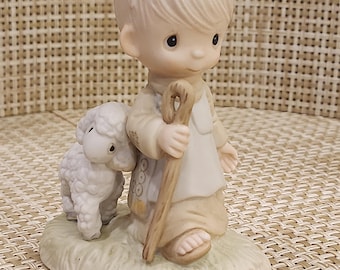 May include: A porcelain figurine depicting a young shepherd with a staff, accompanied by a lamb. The boy is dressed in a beige robe and has fair hair. The lamb is white and fluffy. The base is oval-shaped with green grass.