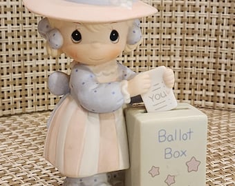 May include: A porcelain figurine of a child wearing a peach-colored hat with a blue ribbon and a matching dress with blue polka dots. The child is placing a white paper slip labeled You into a rectangular ballot box marked Ballot Box with stars.