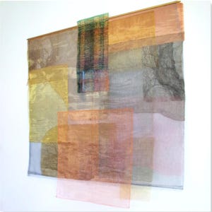 Puede incluir: Un tapiz de pared compuesto por paneles de tela transparente y superpuesta en tonos oro, naranja, rosa y gris. Las piezas rectangulares cuelgan de una fina varilla de madera, creando una composición abstracta con texturas superpuestas.