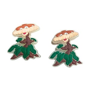 May include: Two mushroom-shaped charms. Each charm features a brown trunk, a beige cap with a brown top, green leaves, and a smiling face with red lips. The charms are set against a white background.