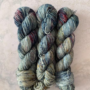May include: Three skeins of hand-dyed yarn in a variegated colorway. The yarn features shades of blue, teal, red, and yellow. The yarn is tightly wound and ready for knitting or crocheting projects. The skeins are displayed on a light gray surface.