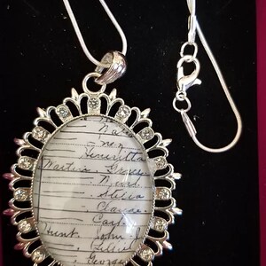 May include: Silver-toned pendant necklace with an oval-shaped pendant. The pendant has a clear center with handwritten text and is surrounded by a decorative silver frame with clear crystals. The necklace is on a black surface.