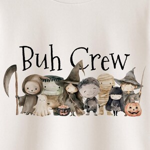 Op de afbeelding: Wit T-shirt met de tekst "Buh Crew" boven een aquarel illustratie van Halloween-personages, waaronder een Grim Reaper, een spook, een heks en een mummie. Het T-shirt hangt aan een houten hanger.